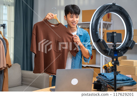 Young online seller man showing his shirt product during online live streaming from his home studio 117629201