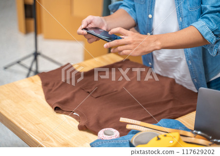 Young online seller man using his phone to take pictures of his shirt product to send to his customers from his home studio Young online seller man using his phone to take pictures of his shirt product to send to his customers from his home studio 117629202