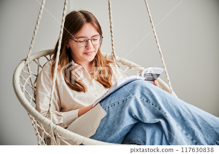 Beautiful girl relaxing and reading book on indoor swing. Pretty female in glasses excited about story she reading. Cozy afternoon with book in hanging chair in home. Beautiful girl relaxing and reading book on indoor swing. Pretty female in glasses excited about story she reading. Cozy afternoon with book in hanging chair in home. 117630838