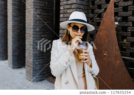 Portrait of successful business woman holding cup of cold drink in hand on her way to work on city street 117635254