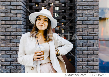 Portrait of successful business woman holding cup of cold drink in hand on her way to work on city street Portrait of successful business woman holding cup of cold drink in hand on her way to work on city street 117635256
