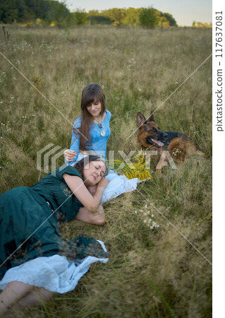 two woman in a whimsical aesthetic style dress with a german shepherd in a field in the summer at sunset 117637081
