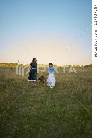 two woman in a whimsical aesthetic style dress with a german shepherd in a field in the summer at sunset 117637187