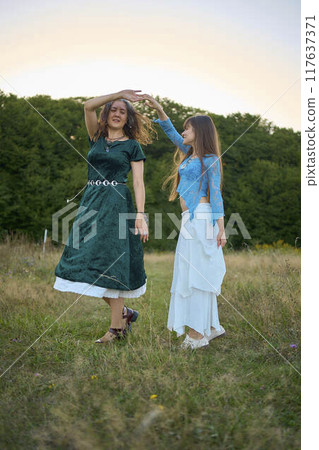 two woman in  whimsical aesthetic style dress  dance in a field in the summer at a sunset 117637371