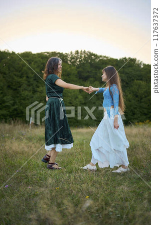 two woman in whimsical aesthetic style dress dance in a field in the summer at a sunset two woman in whimsical aesthetic style dress dance in a field in the summer at a sunset 117637372
