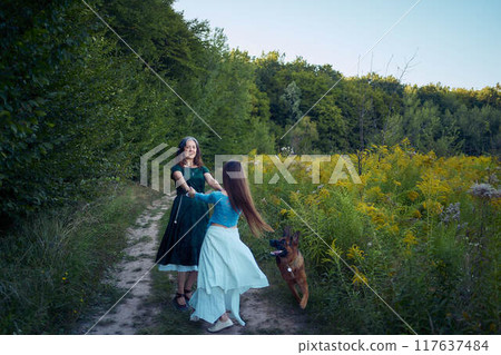 two woman in a whimsical aesthetic style dress with a german shepherd in a field in the summer at sunset 117637484