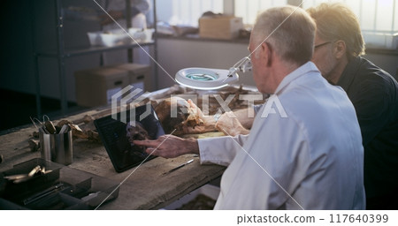 Two scientists examine fossil skull, discuss 3D model displayed on tablet screen Two scientists examine fossil skull, discuss 3D model displayed on tablet screen 117640399
