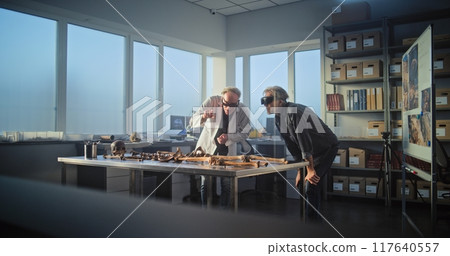 Two archaeologists wearing AR headsets study remains of prehistoric human skeleton Two archaeologists wearing AR headsets study remains of prehistoric human skeleton 117640557