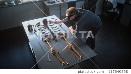 High angle of archaeologist in AR headset studying remains of prehistoric human skeleton 117640568