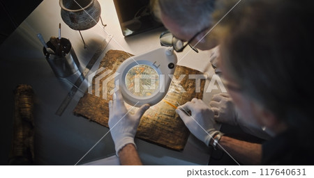 Two archaeologists look through magnifying lamp, study ancient Egyptian papyrus Two archaeologists look through magnifying lamp, study ancient Egyptian papyrus 117640631