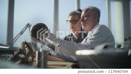 Paleoanthropologist holds prehistoric human skull, archaeologist uses tablet 117640704