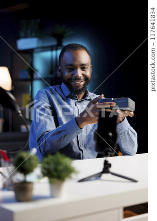 African american internet show host using smartphone on phone holder, filming content about V mount battery. Media star recording rechargeable accumulator review for video production fans 117641834