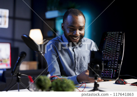 Smartphone on holder capturing footage of excited reviewer discussing with viewers about mechanical keyboard. Online streamer chats with followers during stream, filming computer peripheral review 117641863