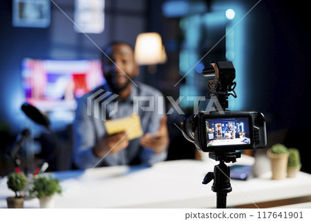 Focus on professional video production camera used for capturing footage of man in blurry background filming literature critique for online readers audience, showcasing new book from publisher Focus on professional video production camera used for capturing footage of man in blurry background filming literature critique for online readers audience, showcasing new book from publisher 117641901