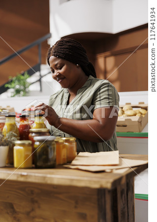 Female consumer searches for fresh, eco friendly produce in sustainable supermarket, supporting local businesses. Black woman checking and opening glass container filled with organic bio product. 117641934