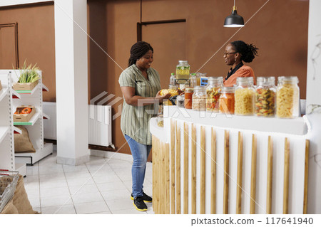 African American woman engages in conversation about environmentally conscious shopping with store attendant at payment counter. Client handing over jars of cereal and honey to vendor for scanning. 117641940