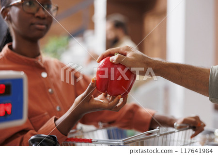 A black woman hands a fresh red bell pepper to a shopkeeper to weigh. African American customer checks the weight of locally grown fruits and vegetables at the cashier's desk. A black woman hands a fresh red bell pepper to a shopkeeper to weigh. African American customer checks the weight of locally grown fruits and vegetables at the cashier's desk. 117641984