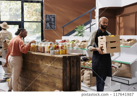 Storekeeper carrying delivered crates full of freshly harvested produce for inventory in eco friendly store. Young male vendor grasping boxes of organic products while customer browses the shop. 117641987