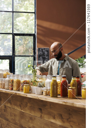 Male shopper glancing around store shelf with variety of freshly harvested products in environmentally friendly packaging. Arab man with beard goes organic shopping to maintain healthy lifestyle. Male shopper glancing around store shelf with variety of freshly harvested products in environmentally friendly packaging. Arab man with beard goes organic shopping to maintain healthy lifestyle. 117641989