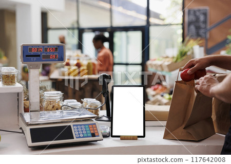 Digital tablet with a white screen display is placed vertically on counter next to a weighing scale. Electronic device showing isolated mockup template while the vendor packs food products in a bag. Digital tablet with a white screen display is placed vertically on counter next to a weighing scale. Electronic device showing isolated mockup template while the vendor packs food products in a bag. 117642008