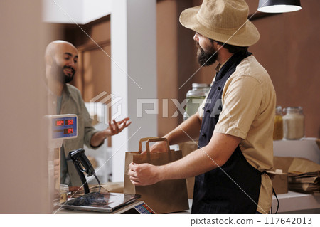 Middle eastern man interacting with caucasian vendor while waiting for his grocery package at checkout counter. Male vendor is packing organic bio food products in paper bag while customer waits. 117642013
