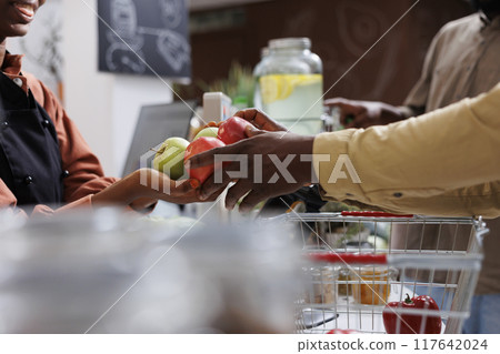 Close-up shot of hands of two african american people holding a bunch of apples over the checkout counter. Selective focus on female vendor receiving fruits from customer for weighing. Close-up shot of hands of two african american people holding a bunch of apples over the checkout counter. Selective focus on female vendor receiving fruits from customer for weighing. 117642024