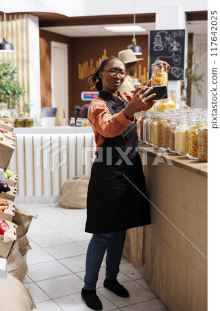 Young black woman films a vlog in an eco store promoting organic, nutritious products. Connects with customers through cell phone for personalized shopping experience. Ideal for marketing campaigns. Young black woman films a vlog in an eco store promoting organic, nutritious products. Connects with customers through cell phone for personalized shopping experience. Ideal for marketing campaigns. 117642025
