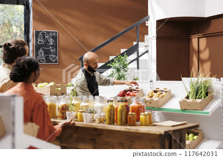 Middle Eastern storekeeper putting a box filled with red apples on shelf in eco friendly store. Male vendor arranges aisle of organic fresh products while two customers, male and female, look on. 117642031