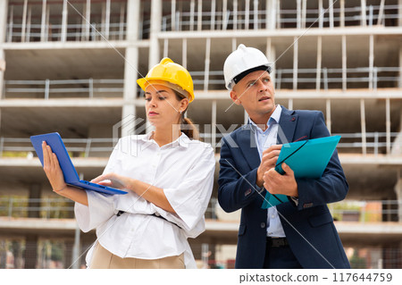 Two architects monitor the progress of work on the construction site, holding a laptop in their hands Two architects monitor the progress of work on the construction site, holding a laptop in their hands 117644759