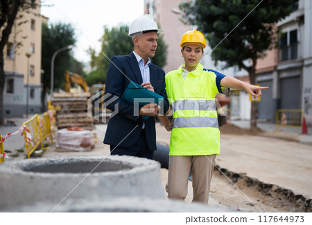 Young woman worker on a construction site shows something to a man engineer Young woman worker on a construction site shows something to a man engineer 117644973