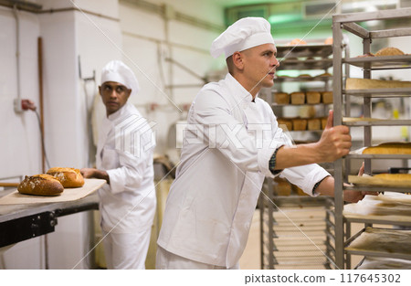 Male baker carrying rack trolley with bread Male baker carrying rack trolley with bread 117645302