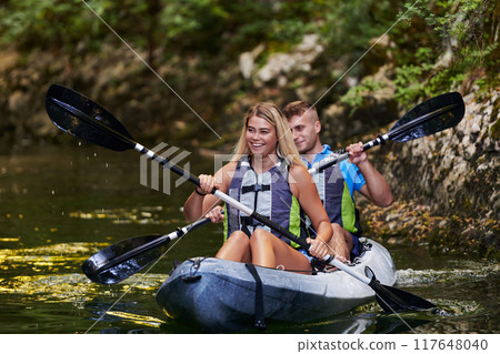 A young couple enjoying an idyllic kayak ride in the middle of a beautiful river surrounded by forest greenery A young couple enjoying an idyllic kayak ride in the middle of a beautiful river surrounded by forest greenery 117648040