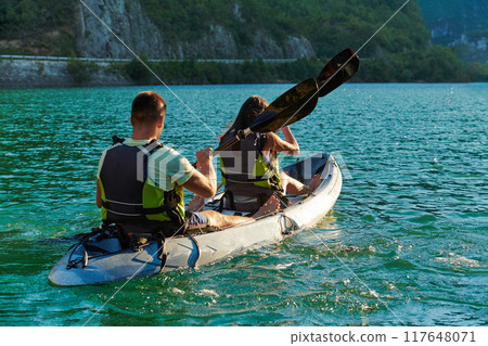 A young couple enjoying an idyllic kayak ride in the middle of a beautiful river surrounded by forest greenery in sunset time  117648071