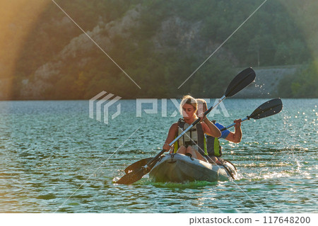 A young couple enjoying an idyllic kayak ride in the middle of a beautiful river surrounded by forest greenery in sunset time  117648200