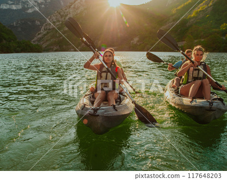 A group of friends enjoying fun and kayaking exploring the calm river, surrounding forest and large natural river canyons during an idyllic sunset. 117648203