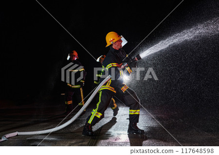 Firefighters using a water hose to eliminate a fire hazard. Team of female and male firemen in dangerous rescue mission. Firefighters using a water hose to eliminate a fire hazard. Team of female and male firemen in dangerous rescue mission. 117648598
