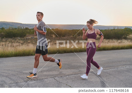A handsome young couple running together during the early morning hours, with the mesmerizing sunrise casting a warm glow, symbolizing their shared love and vitality A handsome young couple running together during the early morning hours, with the mesmerizing sunrise casting a warm glow, symbolizing their shared love and vitality 117648616