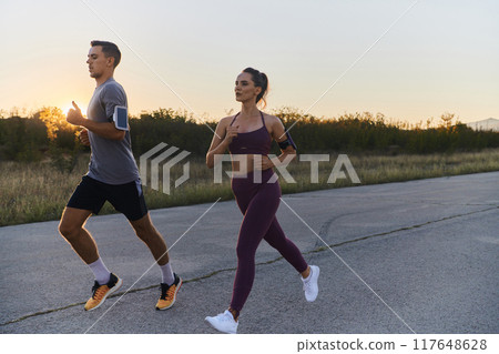 A handsome young couple running together during the early morning hours, with the mesmerizing sunrise casting a warm glow, symbolizing their shared love and vitality 117648628
