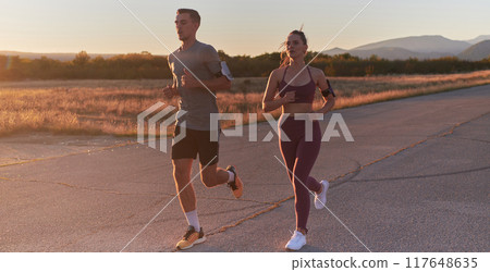 A handsome young couple running together during the early morning hours, with the mesmerizing sunrise casting a warm glow, symbolizing their shared love and vitality A handsome young couple running together during the early morning hours, with the mesmerizing sunrise casting a warm glow, symbolizing their shared love and vitality 117648635