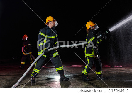 Firefighters using a water hose to eliminate a fire hazard. Team of female and male firemen in dangerous rescue mission.  117648645