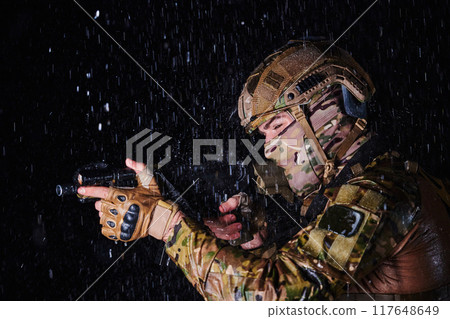 Army soldier in Combat Uniforms with an assault rifle, plate carrier and combat helmet going on a dangerous mission on a rainy night. Army soldier in Combat Uniforms with an assault rifle, plate carrier and combat helmet going on a dangerous mission on a rainy night. 117648649