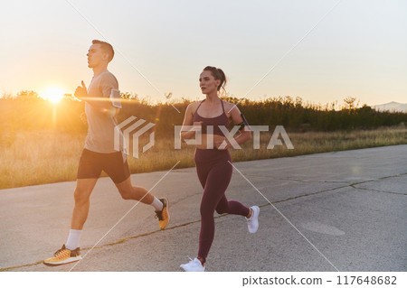 A handsome young couple running together during the early morning hours, with the mesmerizing sunrise casting a warm glow, symbolizing their shared love and vitality 117648682