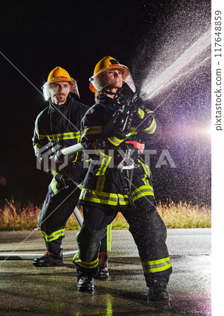 Firefighters using a water hose to eliminate a fire hazard. Team of female and male firemen in dangerous rescue mission. Firefighters using a water hose to eliminate a fire hazard. Team of female and male firemen in dangerous rescue mission. 117648859
