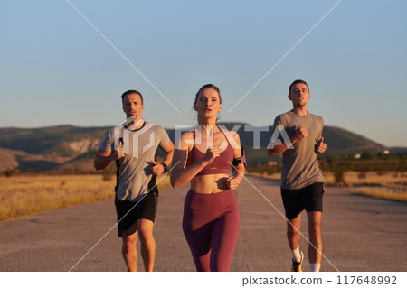 A group of young athletes running together in the early morning light of the sunrise, showcasing their collective energy, determination, and unity A group of young athletes running together in the early morning light of the sunrise, showcasing their collective energy, determination, and unity 117648992