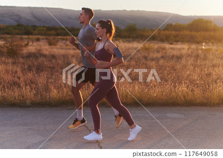 A handsome young couple running together during the early morning hours, with the mesmerizing sunrise casting a warm glow, symbolizing their shared love and vitality A handsome young couple running together during the early morning hours, with the mesmerizing sunrise casting a warm glow, symbolizing their shared love and vitality 117649058