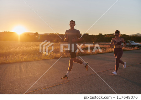 A handsome young couple running together during the early morning hours, with the mesmerizing sunrise casting a warm glow, symbolizing their shared love and vitality A handsome young couple running together during the early morning hours, with the mesmerizing sunrise casting a warm glow, symbolizing their shared love and vitality 117649076