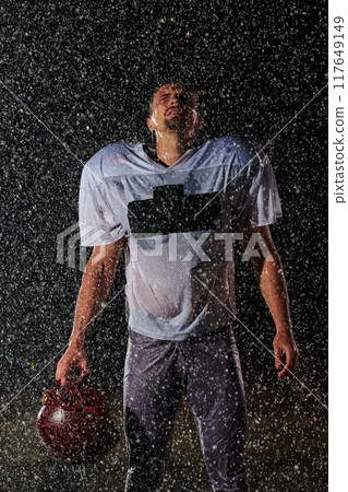 American Football Field: Lonely Athlete Warrior Standing on a Field Holds his Helmet and Ready to Play. Player Preparing to Run, Attack and Score Touchdown. Rainy Night with Dramatic Fog, Blue Light 117649149