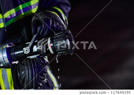 Firefighter using a water hose to eliminate a fire hazard. Team of firemen in the dangerous rescue mission. Firefighter using a water hose to eliminate a fire hazard. Team of firemen in the dangerous rescue mission. 117649195