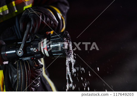 Firefighter using a water hose to eliminate a fire hazard. Team of firemen in the dangerous rescue mission. Firefighter using a water hose to eliminate a fire hazard. Team of firemen in the dangerous rescue mission. 117649203