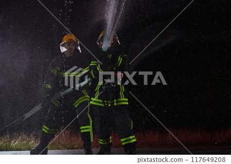 Firefighters using a water hose to eliminate a fire hazard. Team of female and male firemen in dangerous rescue mission. Firefighters using a water hose to eliminate a fire hazard. Team of female and male firemen in dangerous rescue mission. 117649208